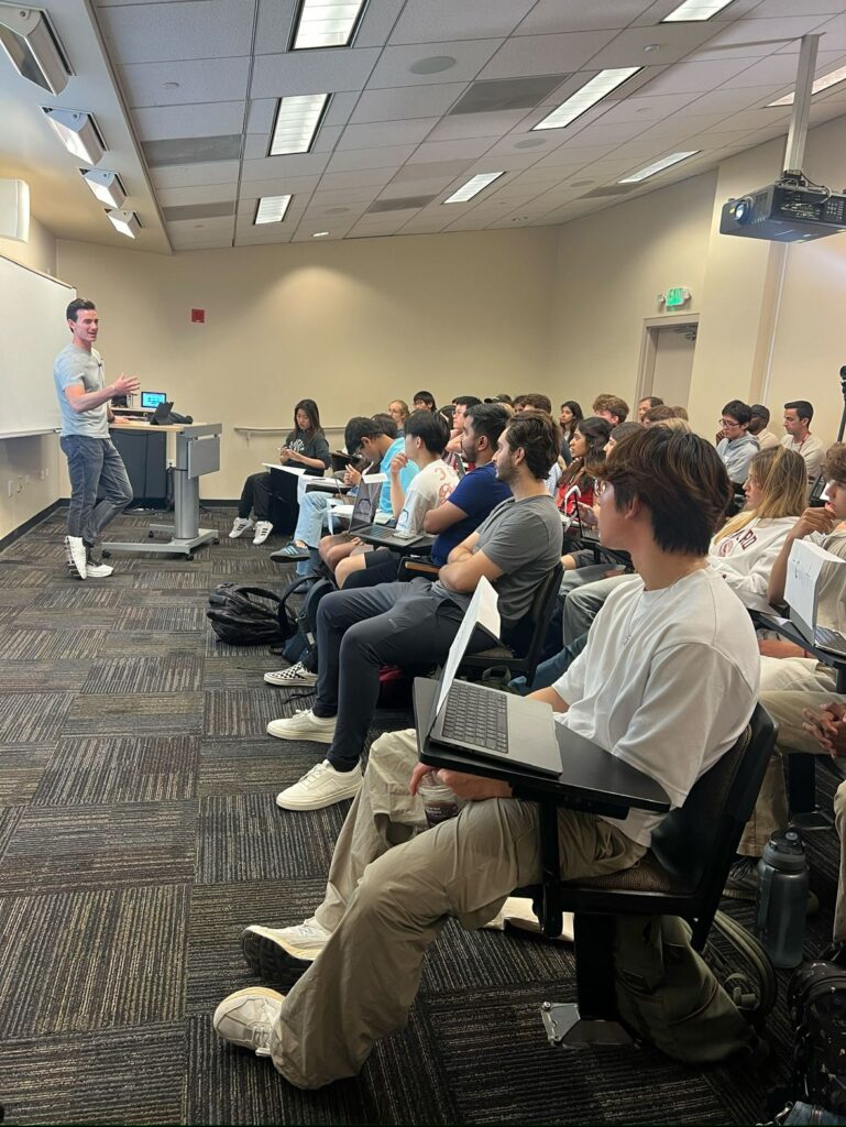Noah Berkson speaking to a room of seated students with laptops in a university classroom